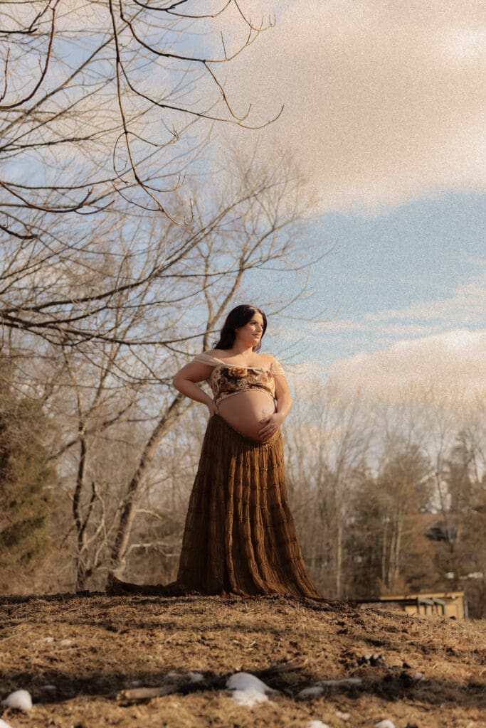 Pregnant woman standing outdoors holding her baby bump during a golden hour maternity photoshoot.