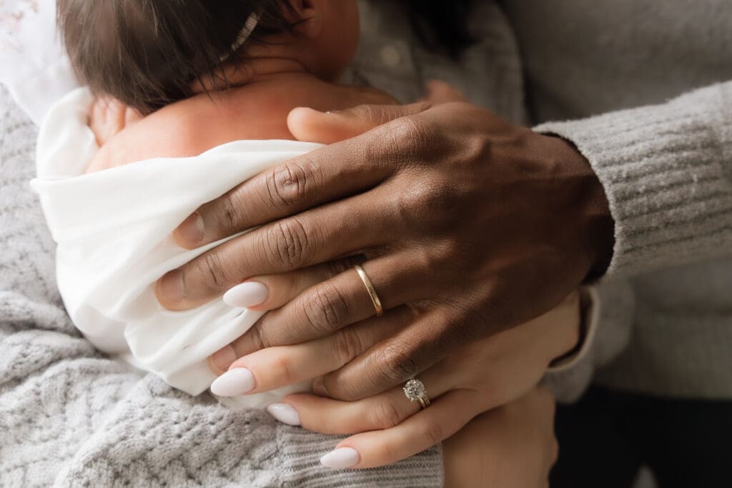 Mother and father have their hands intertwined on newborn baby's back holding baby with love and calmness. 